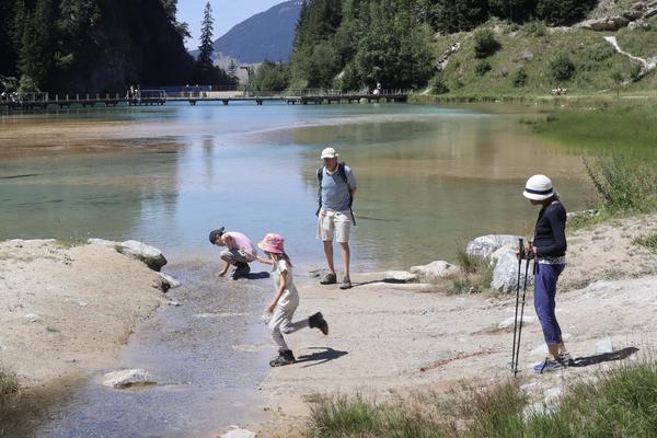 A family enjoys a sunny day by a clear, shallow lake in a mountainous landscape, with two children playfully interacting with the water at the shore